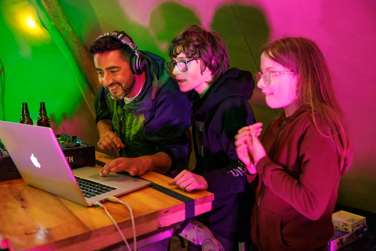 young people and adult DJ on laptop in tent, coloured lights
