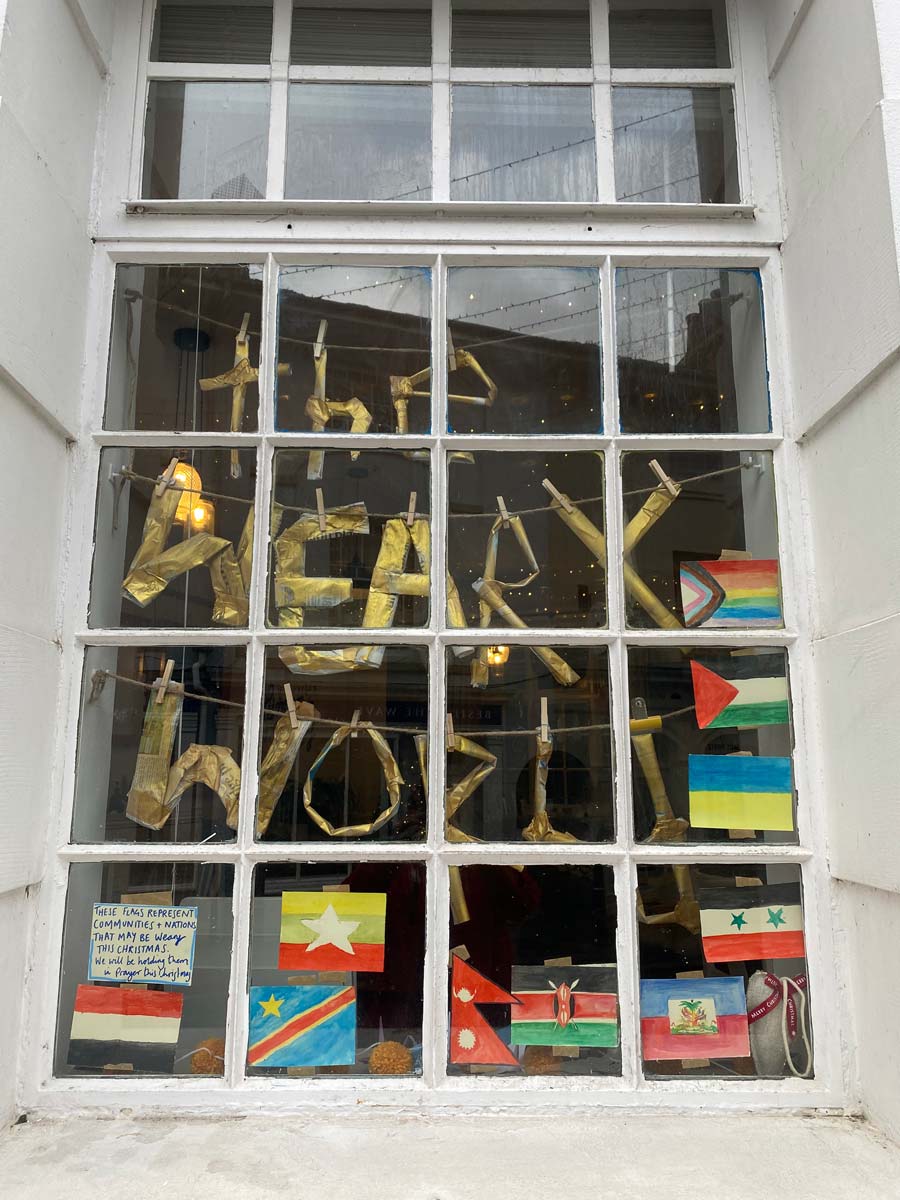 Window display seen from street with letters spelling The Weary World hung with clothes pegs and flags of various nations, with promise to pray