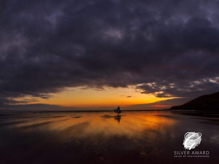 lone paddleboarder walsk across a beach at sunset with heavy dark clouds dominating; Silver Award logo from Guild of Photographers
